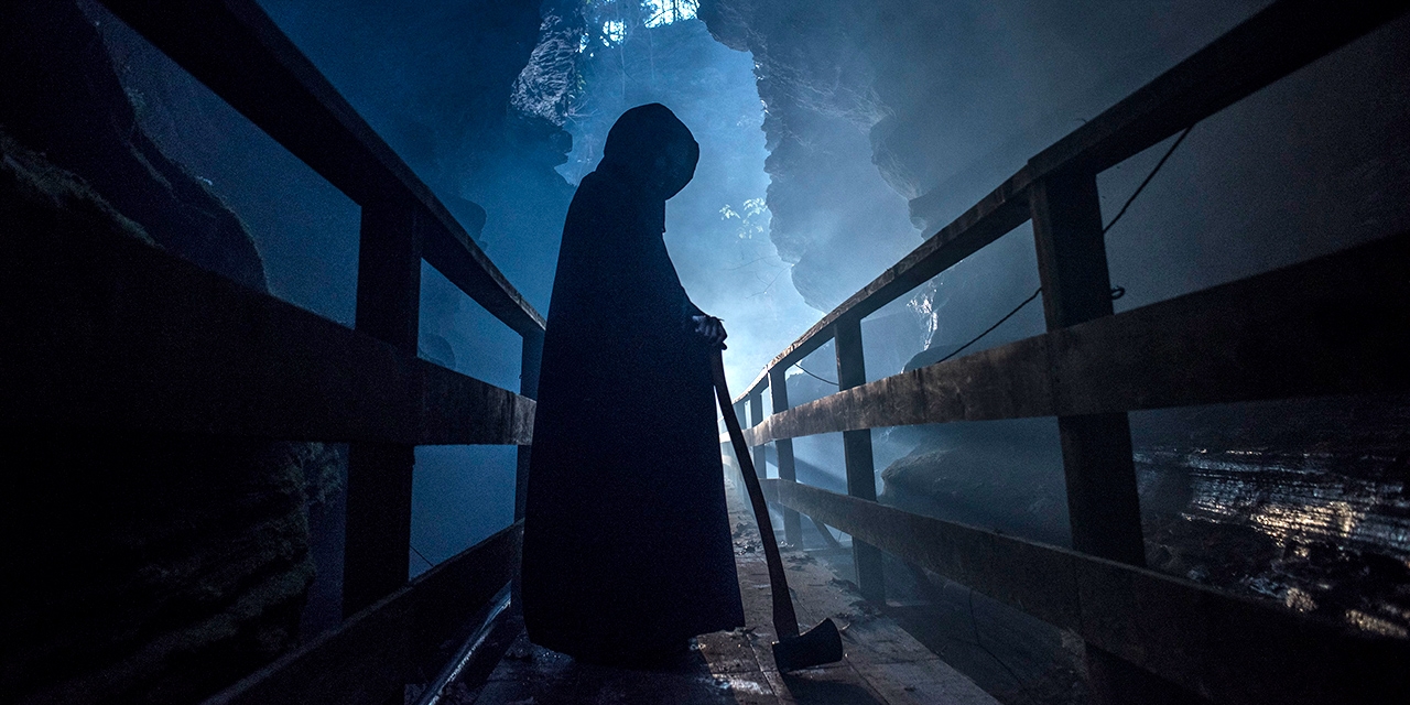 A hooded figure stands silhouetted with an axe in a spooky gulch.