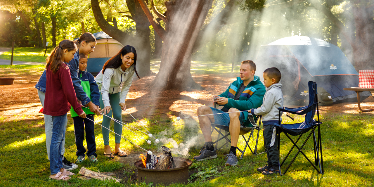 A family sits around a fire pit roasting marshmallows with friends playing lawn games and socializing in the background with tents and a camper behind them in Wisconsin Dells.