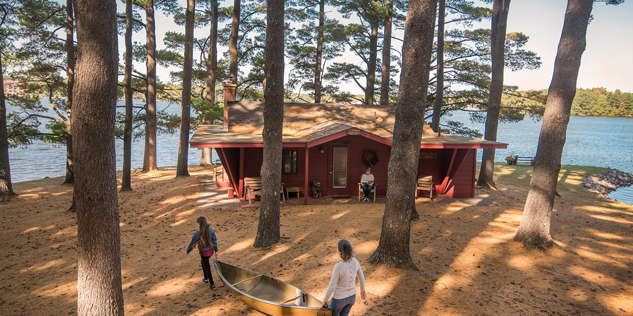 A cabin on a peninsula with people carrying a canoe to the lake.