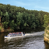 A Dells Boat Tours boat sails past rock cliffs and woods.