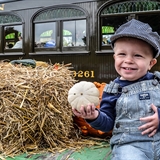 A child holding a small pumpkin in front of a train car.