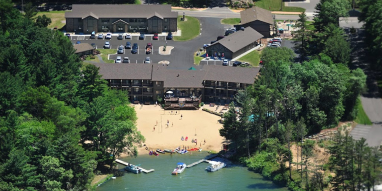 Outdoor pool area with beach, volleyball net, waterfront, and wooded area.