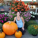 A woman poses in front of flowers and large pumpkins.